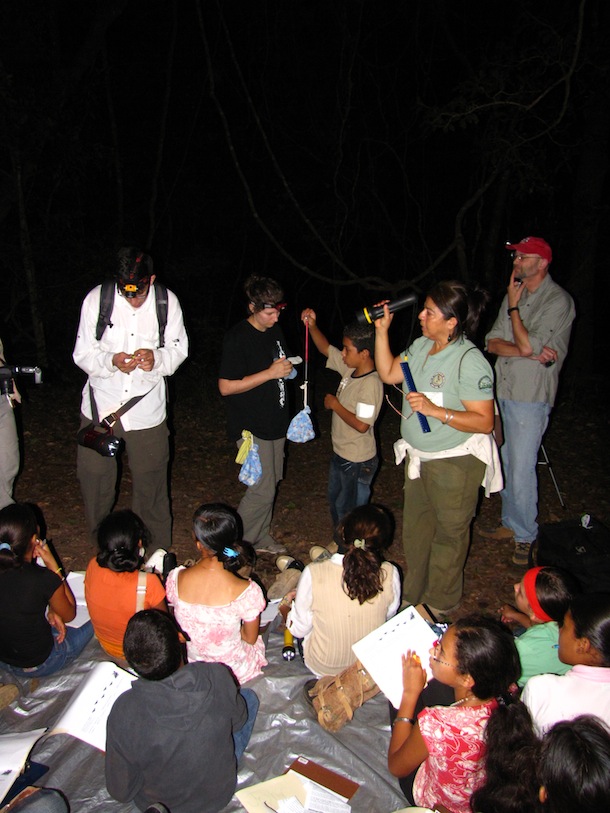 At night, with flashlights and headlamps, school children and teacher out with bat researcher weighing bat in cloth bag.
